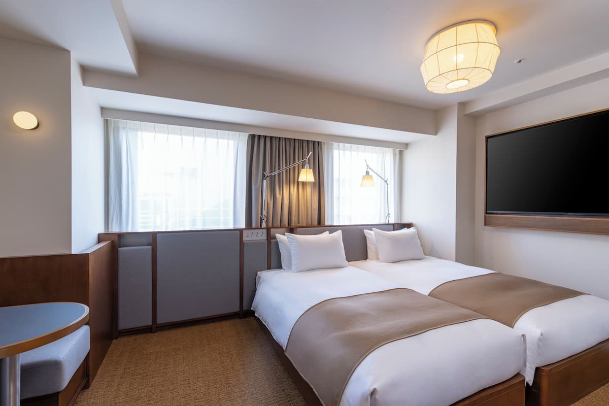 A hotel room with a bed, a desk, a chair, a window with curtains, and a flat-screen TV.