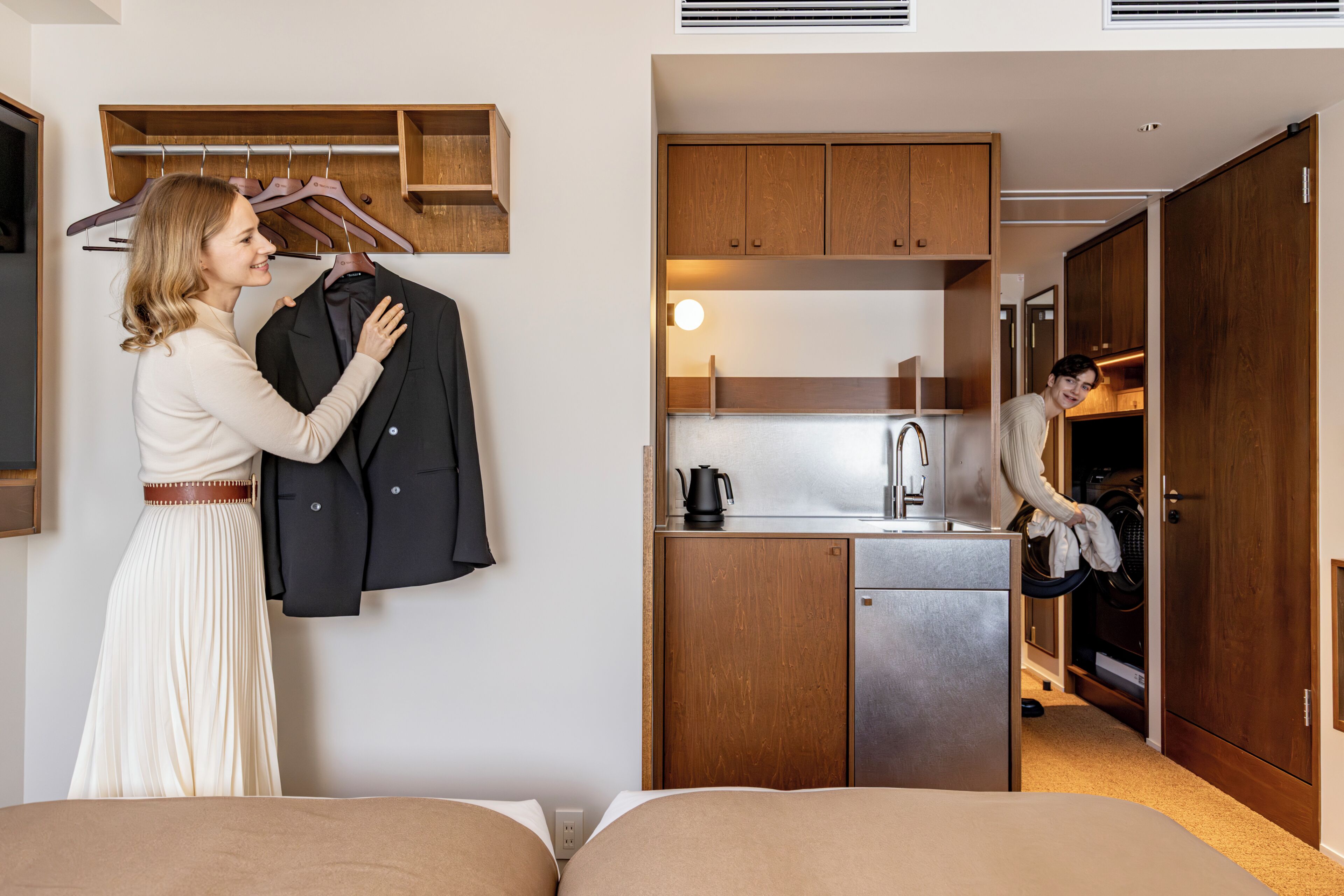 A compact hotel room with a bed, a kitchenette, a wooden wardrobe, and a hanging clothes rack.