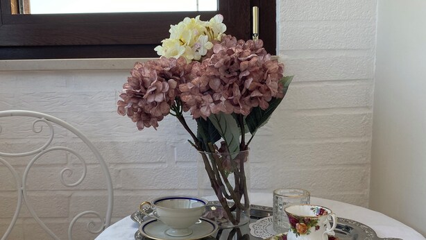 Una mesa con mantel de encaje blanco, bandeja plateada con tazas de té y un florero con flores.