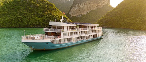 A large riverboat with multiple decks, surrounded by lush greenery and rocky cliffs.