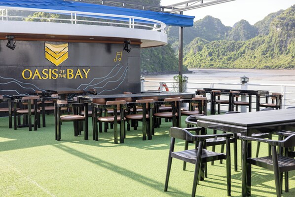 A dining area with tables and chairs on a ship deck, featuring a sign that reads 'OASIS OF THE BAY'. - Oasis Bay Party Cruise (Tuan Chau)