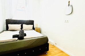 Uma cama com cabeceira escura, duas almofadas, uma toalha enrolada e um violão pendurado na parede.
