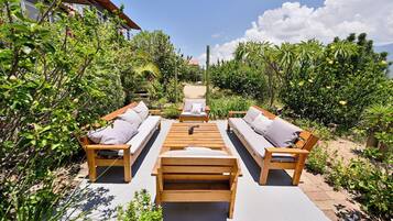 Un área de estar al aire libre con mobiliario de madera y una mesa central, rodeada de abundante vegetación y un cielo despejado.
