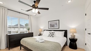 Un dormitorio con cama, ventilador de techo, lámparas de noche, ventana con cortinas, un cuadro en la pared y un aparador oscuro.