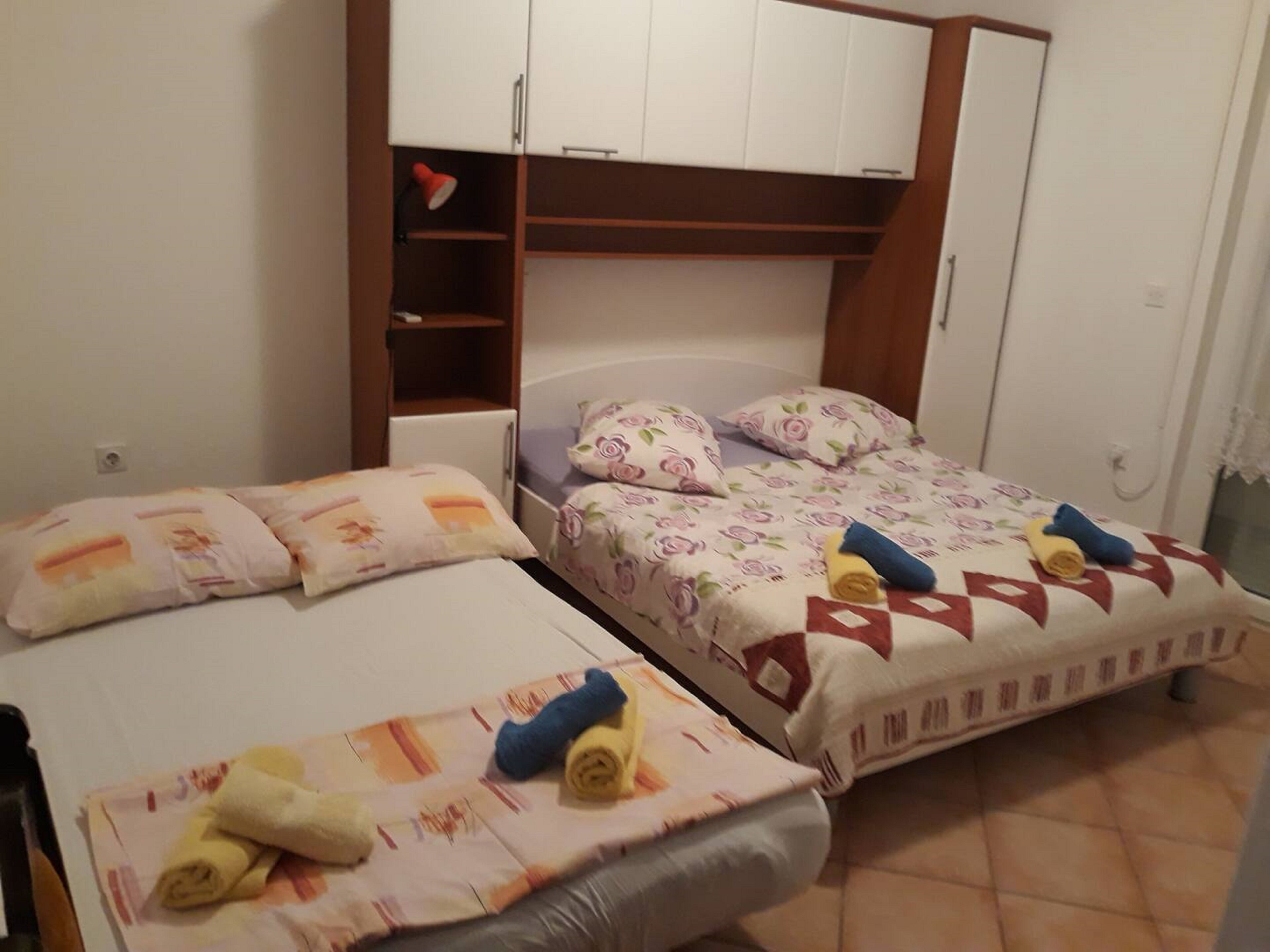 Two single beds with floral bedding, a wooden wardrobe, and a small shelf with a red lamp.