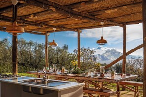 Un espace de restauration en plein air avec une table en bois dressée pour un repas, un évier encastré et des lampes suspendues.