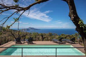 Une piscine rectangulaire bordée de quatre chaises longues sur une terrasse en bois, offrant une vue sur un paysage côtier avec des montagnes et la mer.
