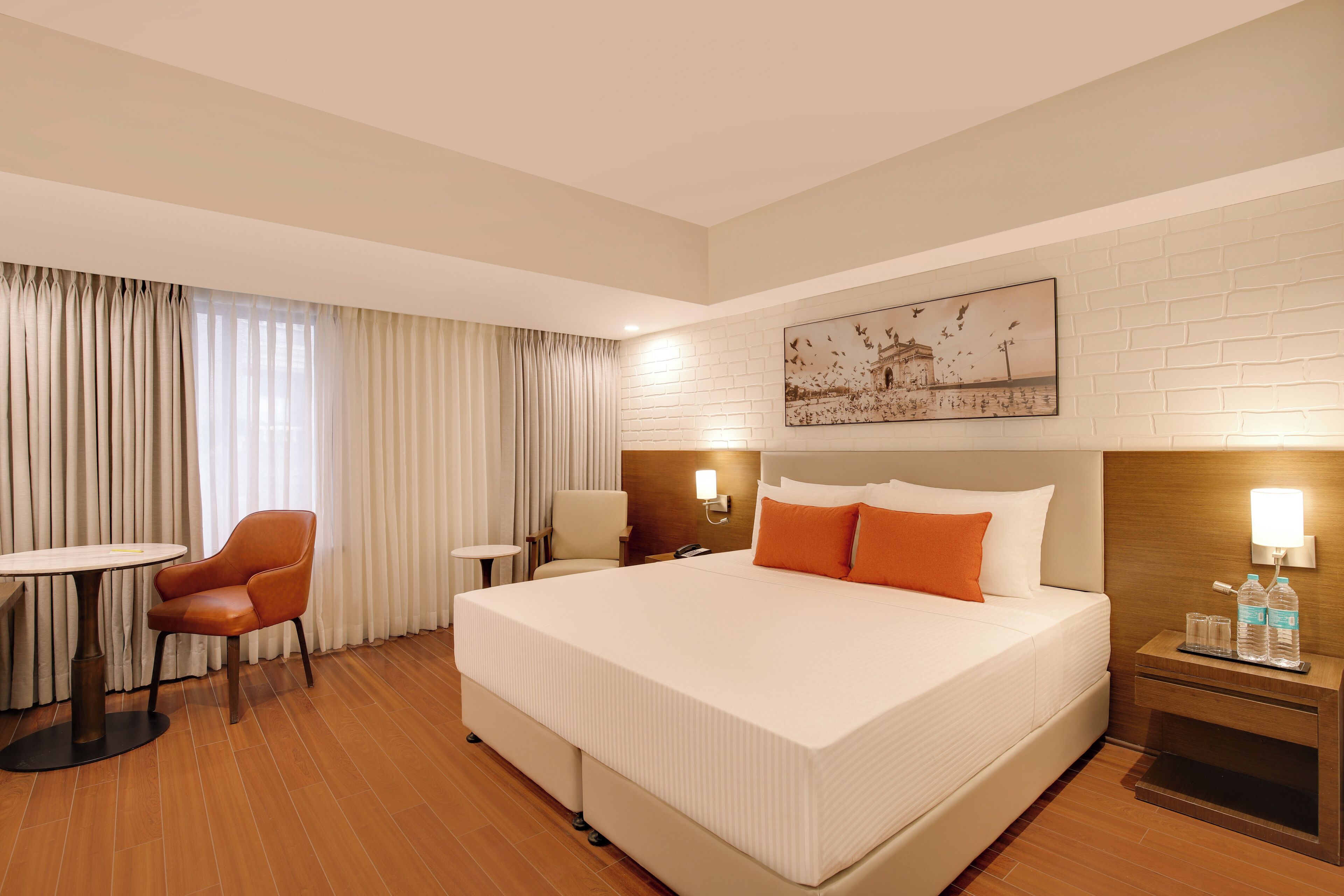 A hotel room with a large bed, a round table, a chair, a nightstand with a lamp, and a wall-mounted artwork.
