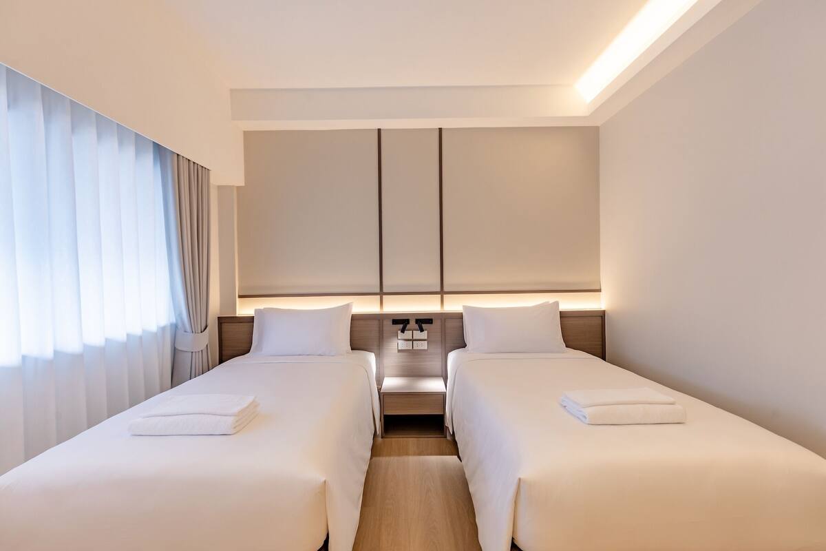Une chambre d’hôtel avec deux lits simples, une table de chevet, un bureau intégré et une grande fenêtre avec des rideaux.