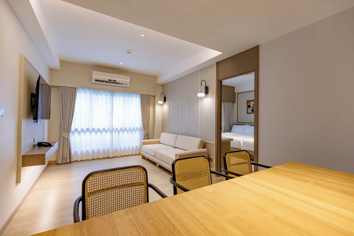Une chambre d’hôtel moderne avec une table à manger en bois, des chaises en osier, un canapé et un téléviseur fixé au mur.