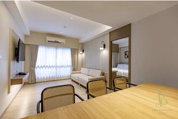 A hotel room with a wooden dining table, two wicker chairs, a sofa, a TV mounted on the wall, and a large mirror.