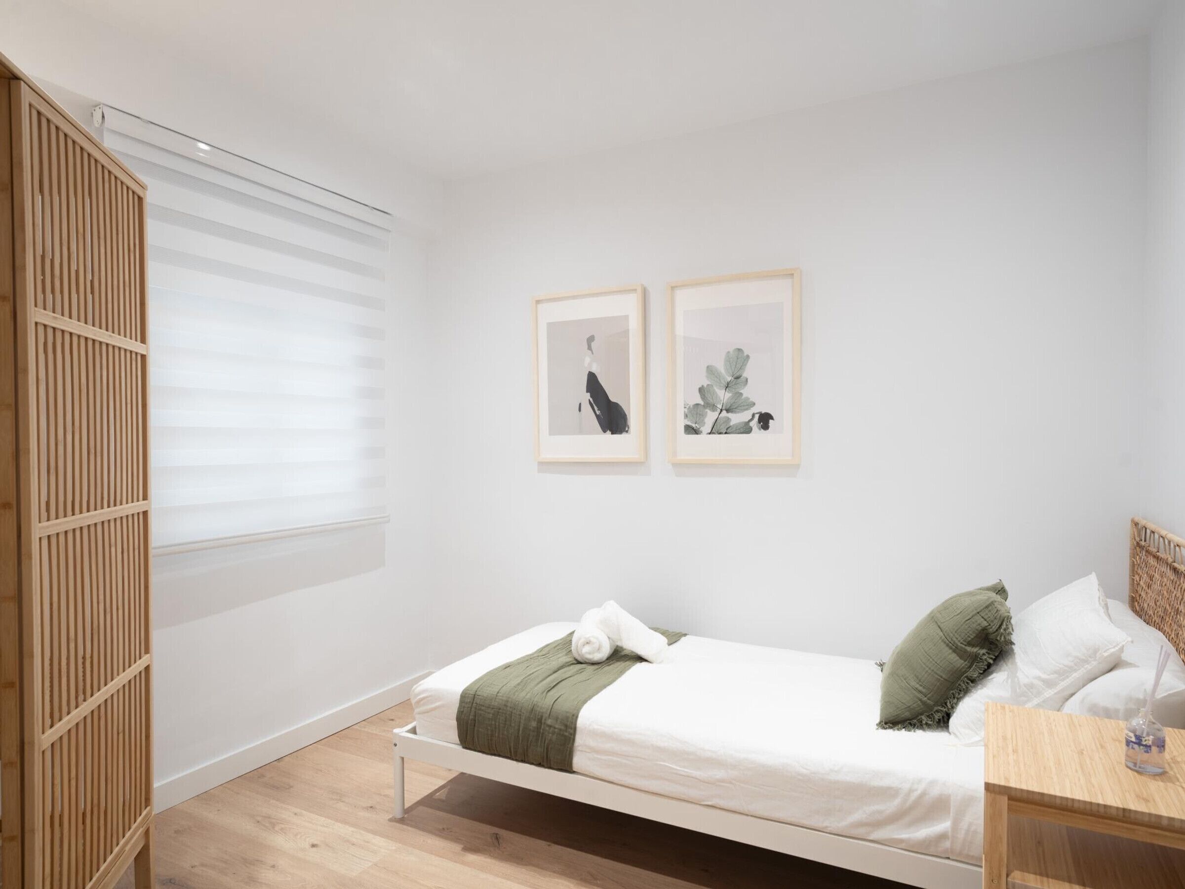 A minimalist bedroom with a single bed, a wooden wardrobe, and two framed botanical prints on the wall.