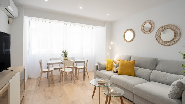 A modern living room with a grey sofa, a dining table with chairs, a TV, and a large window with sheer curtains.