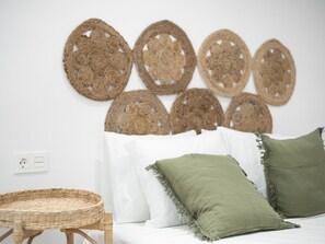 A bedroom with a white bed, two green pillows, a wicker chair, and a wall decorated with woven wall hangings.