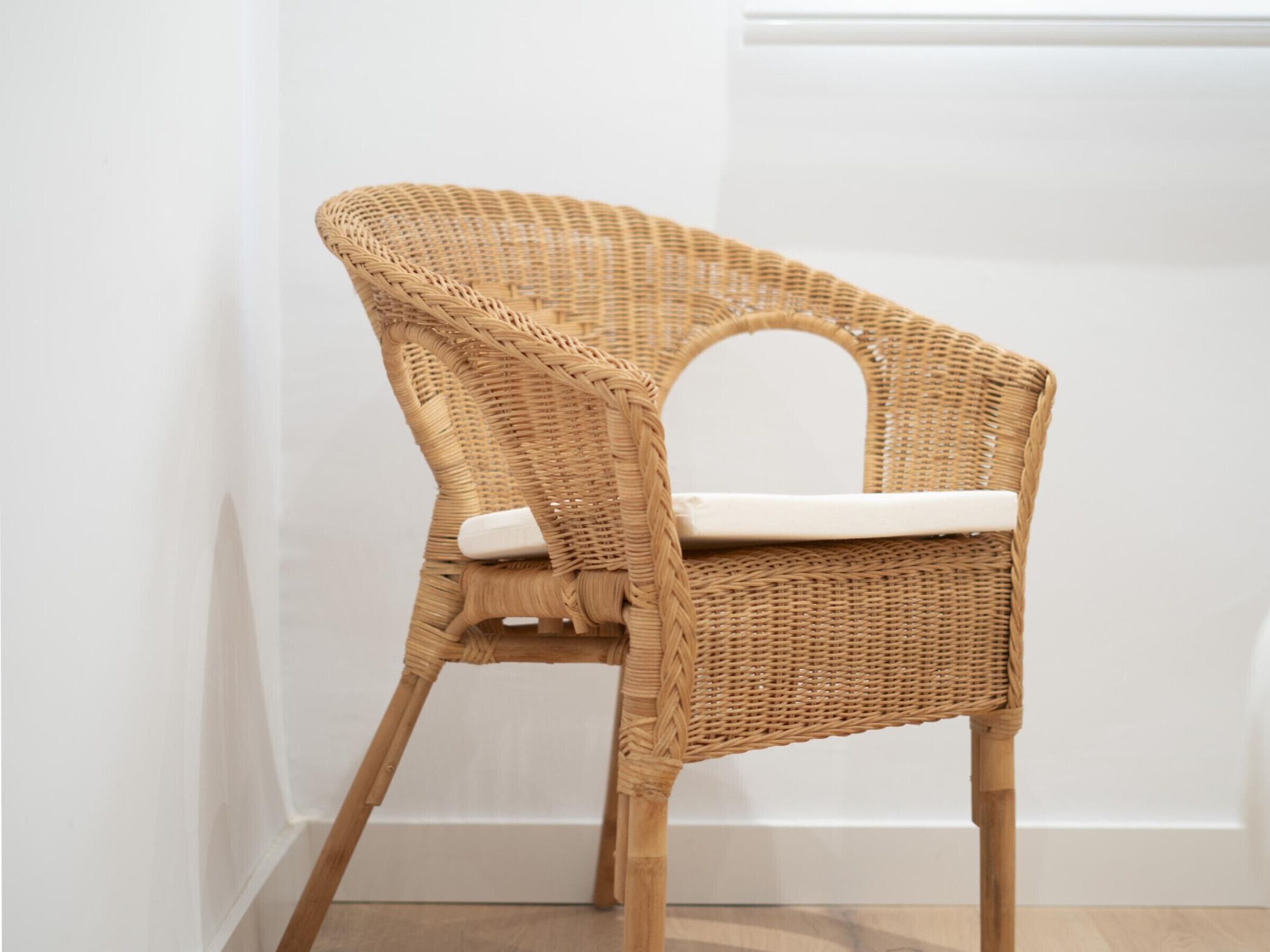 A wicker chair with a white cushion, positioned against a white wall with a wooden floor.
