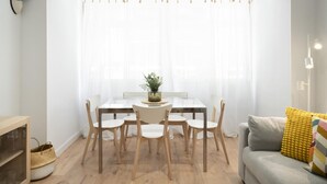 A modern living room with a glass dining table surrounded by wooden chairs, a grey sofa with a yellow cushion, and a large window with sheer curtains.
