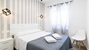 Habitación de hotel moderna con una cama grande, una mesita de noche, una silla y un ventanal con cortinas de gasa.