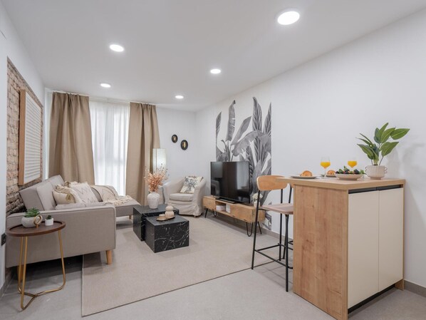 A modern living room with a sofa, coffee table, TV, and a small kitchen area with a bar stool and counter.