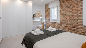 A bedroom with a brick wall, a bed with white linens, a desk with a chair, and a window with a view.
