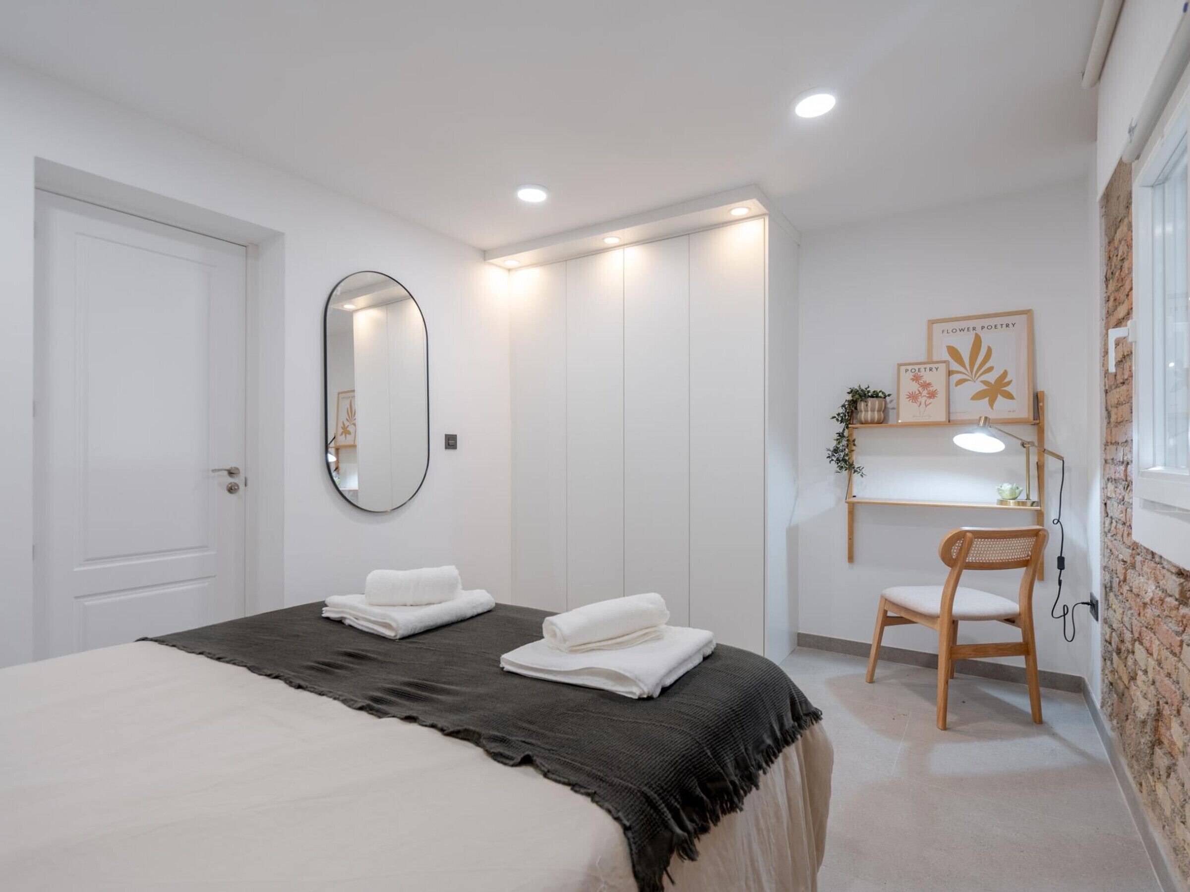 A bedroom with a bed, a mirror, a bedside table with a lamp, a chair, and a wall with a shelf holding decorative items.
