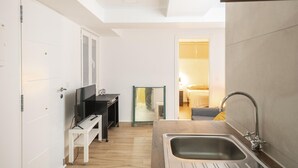 Een kleine keuken met een spoelbak, een eethoekje en een zichtbare slaapkamer met een bed en nachtkastje.