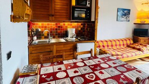 Een functionele keuken met houten kastjes, een magnetron en een koffiezetapparaat. Er is een eettafeltje en een bed met een geruit plaid.