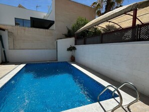 Una piscina rectangular con una escalera metálica, rodeada por un muro de ladrillo blanco y una zona cubierta con cerramiento de metal.