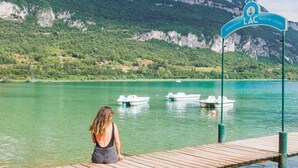 Une femme est assise sur une jetée en bois, d’où elle a une vue sur un lac, des bateaux, une enseigne de restaurant et des montagnes en arrière-plan.