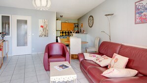 Un salon avec un canapé en cuir rouge, une petite table et un fauteuil. Au fond, on aperçoit une cuisine avec une table à manger et des chaises.