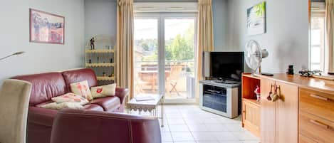 A living room with a leather sofa, a TV, a wooden cabinet, and a balcony with a view of greenery.
