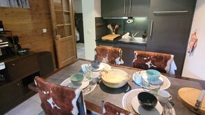 Un coin repas avec une table en bois dressée pour quatre personnes, entourée de chaises recouvertes de cuir de vache. On aperçoit à l’arrière-plan une cuisine avec des meubles sombres et un réfrigérateur en acier inoxydable.