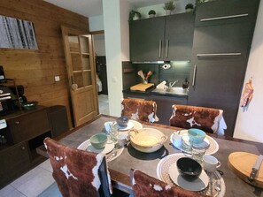 Ein Essbereich mit einem Holztisch, gedeckt für vier Personen, umgeben von Stühlen mit Kuhfellbezug. Im Hintergrund ist eine Küche mit dunklen Schränken und einem Edelstahlkühlschrank zu sehen.