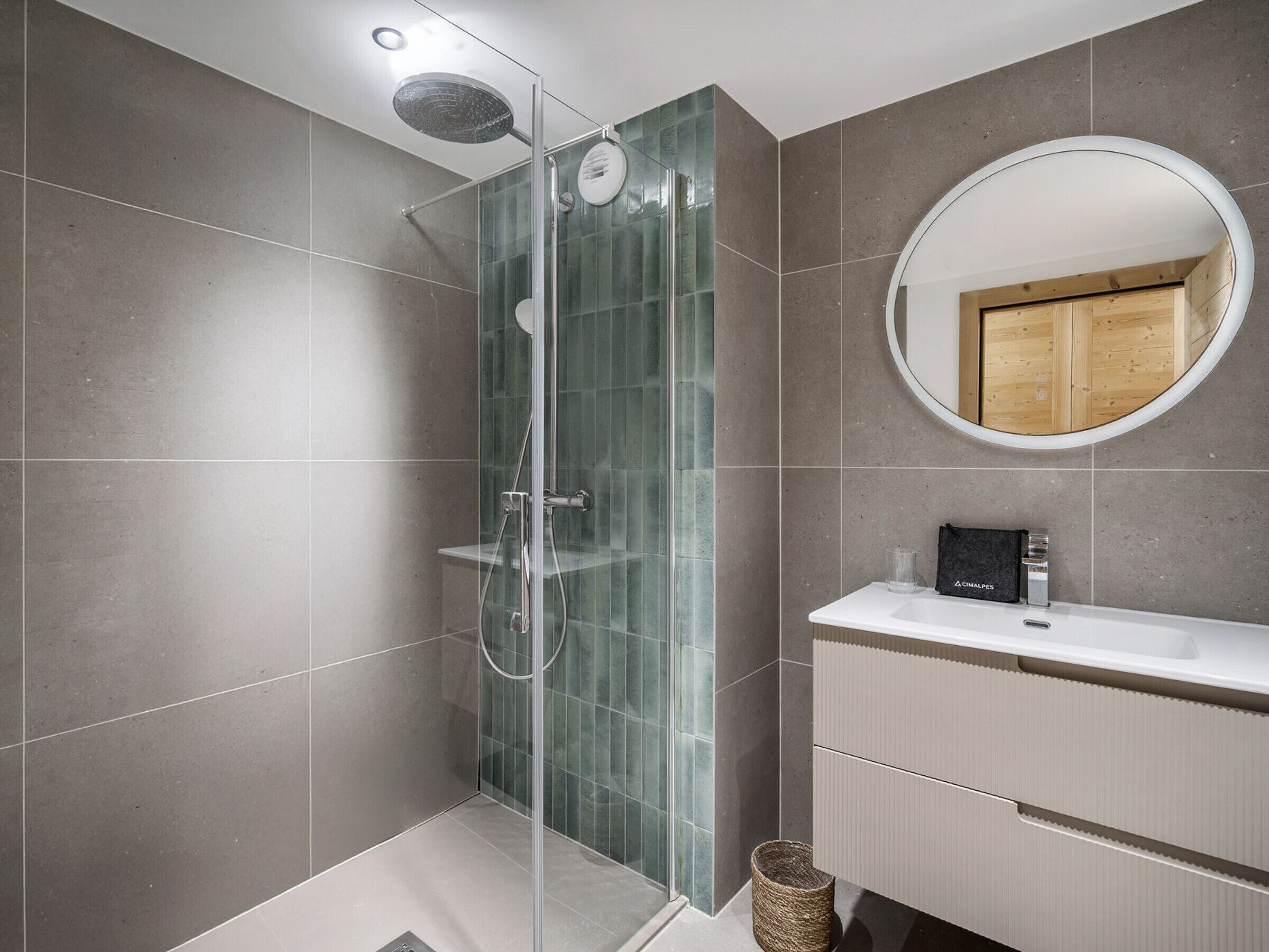 Un baño moderno con ducha envuelta en vidrio, un espejo redondo y un mueble de tocador de madera.