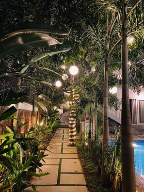 A pathway lined with tropical plants and illuminated by spherical lights, leading to a pool area.