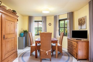 A dining area with a wooden table, wicker chairs, a TV on a wooden cabinet, and a large window with curtains.