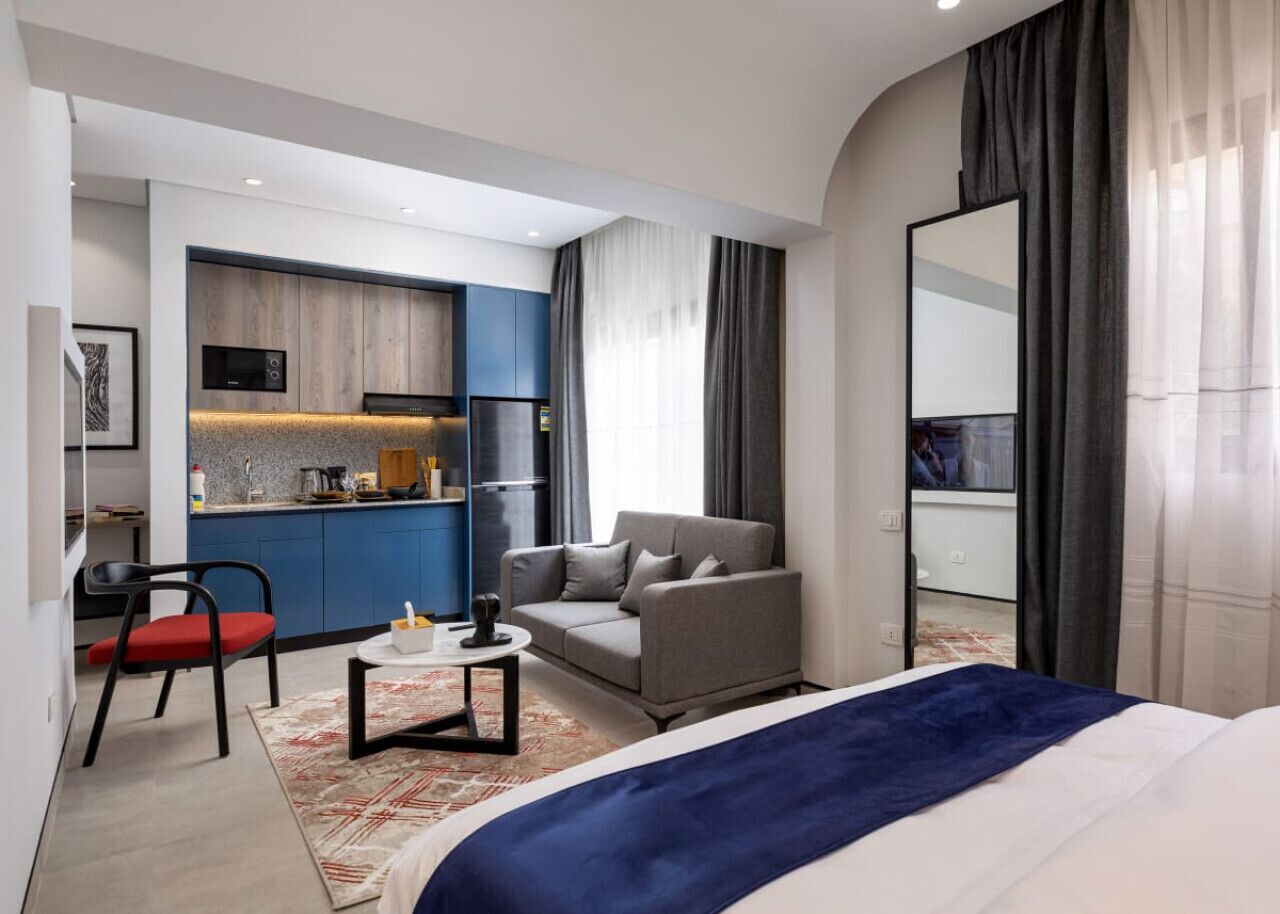 Habitació d'hotel moderna amb un llit gran, un sofà gris, una taula petita i una cuineta al fons.
