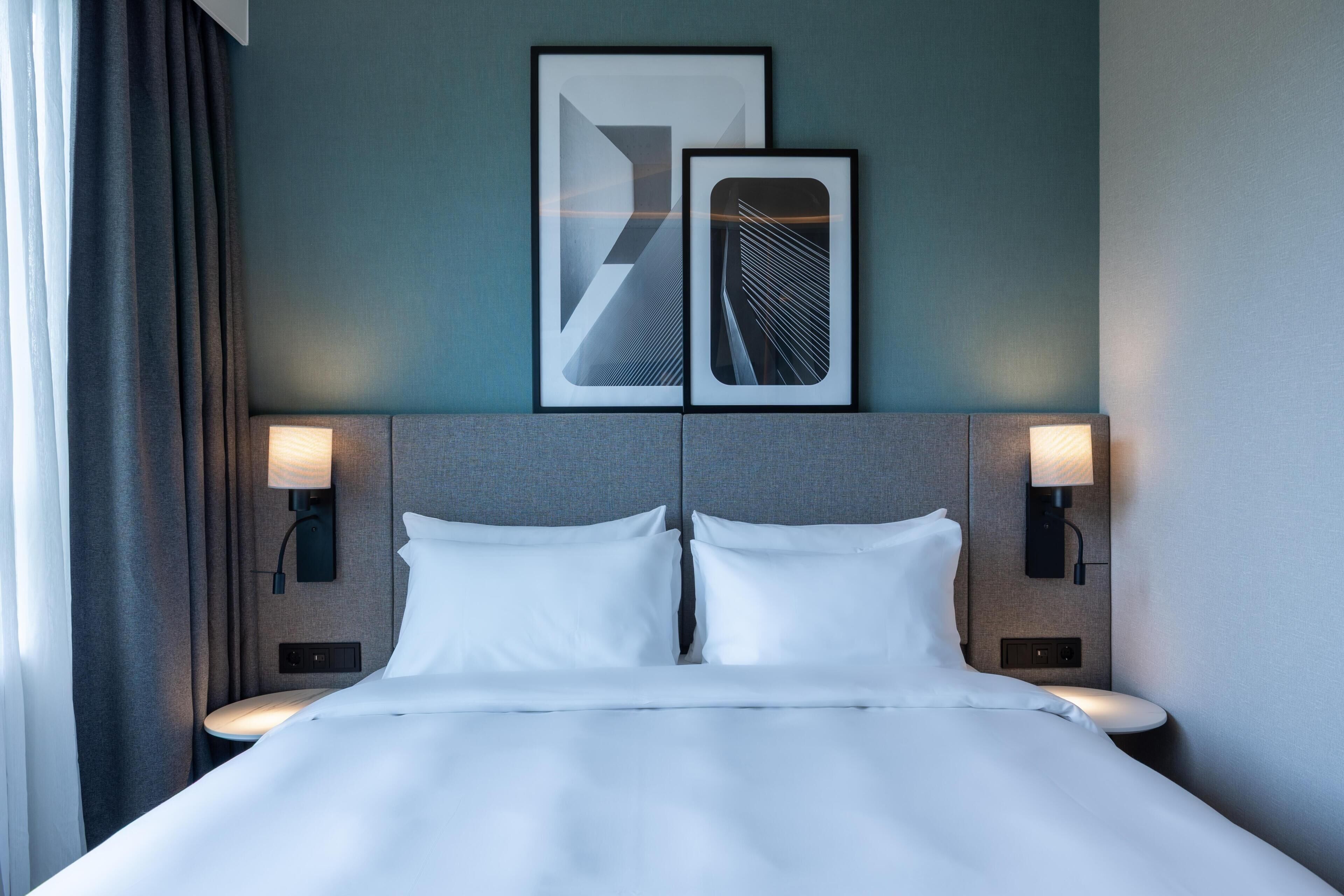 Een hotelkamer met een bed, twee wandlampen en twee abstracte kunstwerken in lijsten aan de muur.
