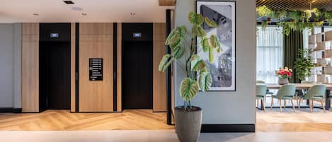 A modern lobby with a reception desk, two elevators, a large potted plant, and a dining area with chairs and a table.