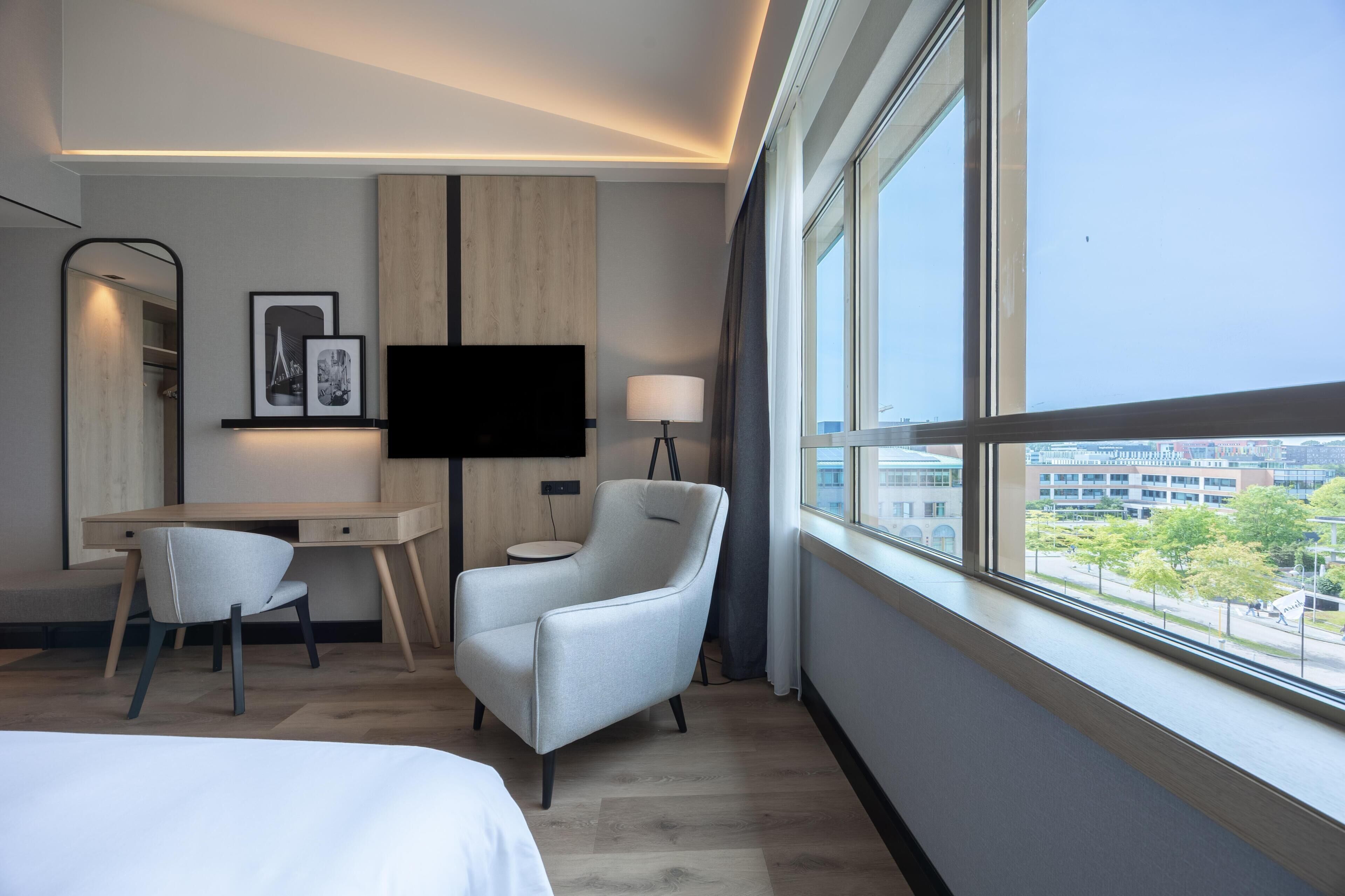 Une chambre d’hôtel moderne équipée d’un bureau, de deux chaises, d’un téléviseur à écran plat, d’une lampe et d’une grande fenêtre donnant sur la ville.