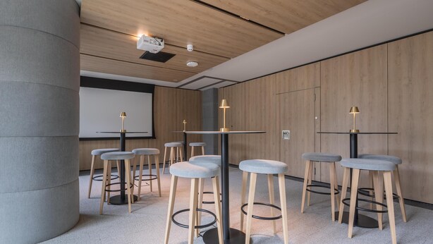 A modern conference room with high tables, grey cushioned stools, and a projector screen. - Radisson Hotel Amsterdam Airport Schiphol Hoofddorp (Hoofddorp)