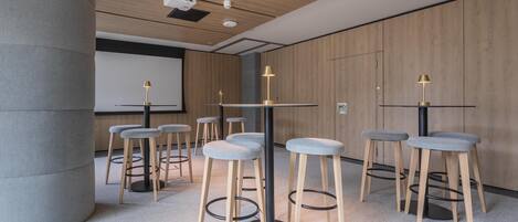 Una sala de conferencias moderna con mesas altas, taburetes acolchados grises y una pantalla de proyector.