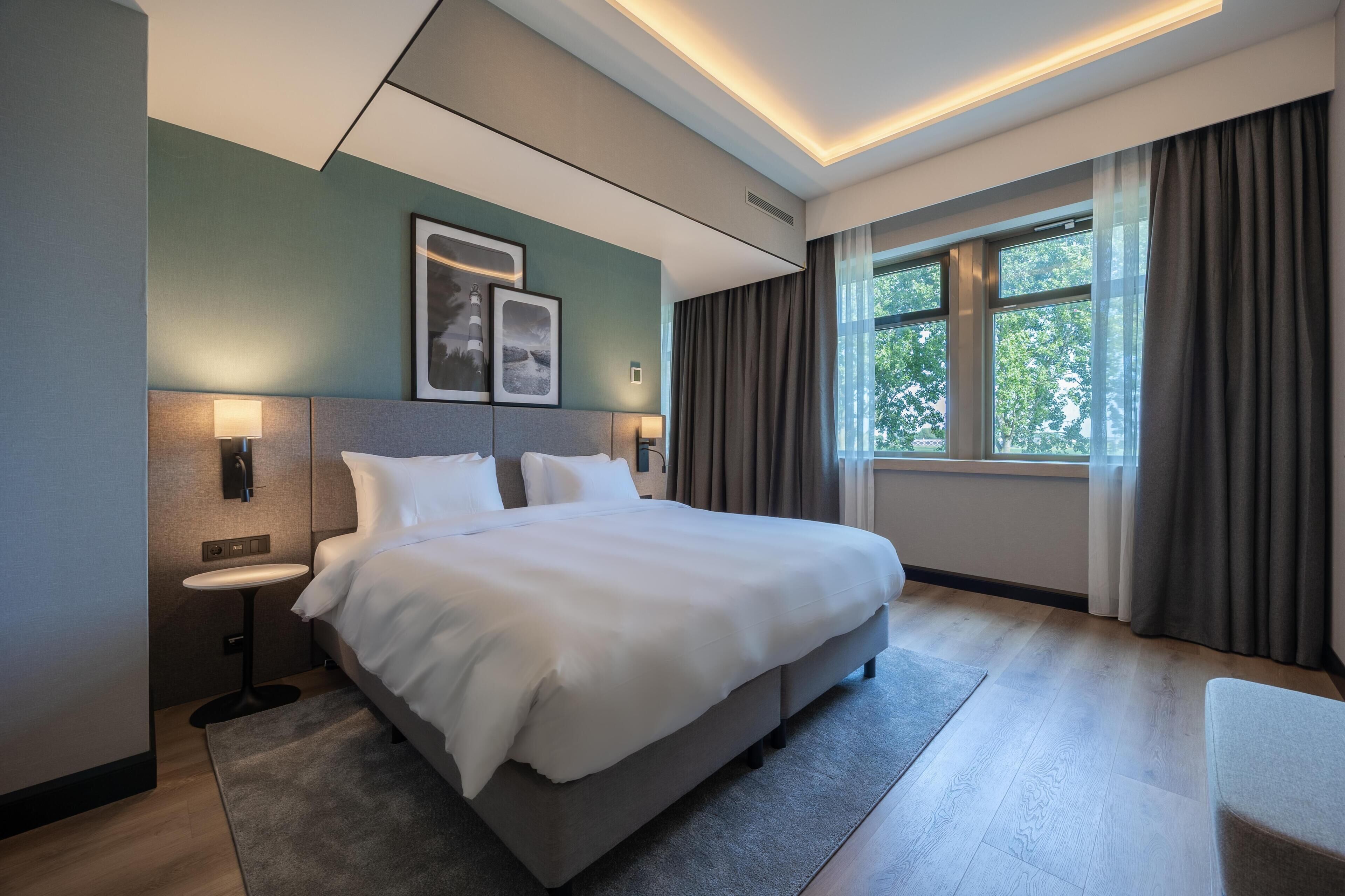 Una habitación de hotel moderna con una cama grande, mesitas de noche, un escritorio pequeño, una ventana con cortinas y cuadros en la pared.