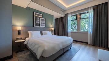 Una habitación de hotel moderna con una cama grande, mesitas de noche, un escritorio pequeño, una ventana con cortinas y cuadros en la pared.