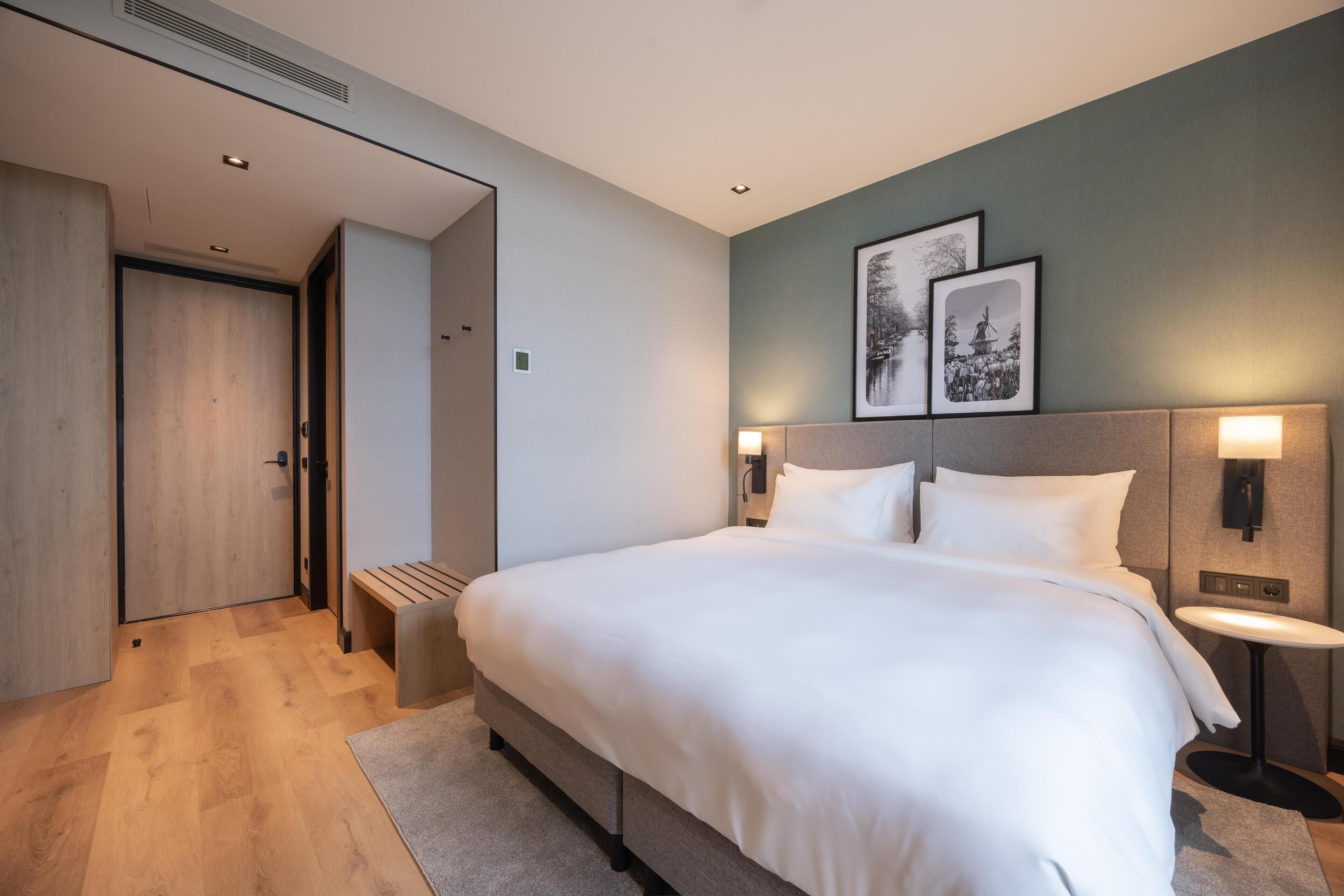 A modern hotel room with a large bed, bedside tables, a bench, and framed pictures on the wall.