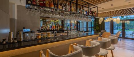 Zona de bar con mostrador, taburetes, estantes con botellas y copas colgantes.