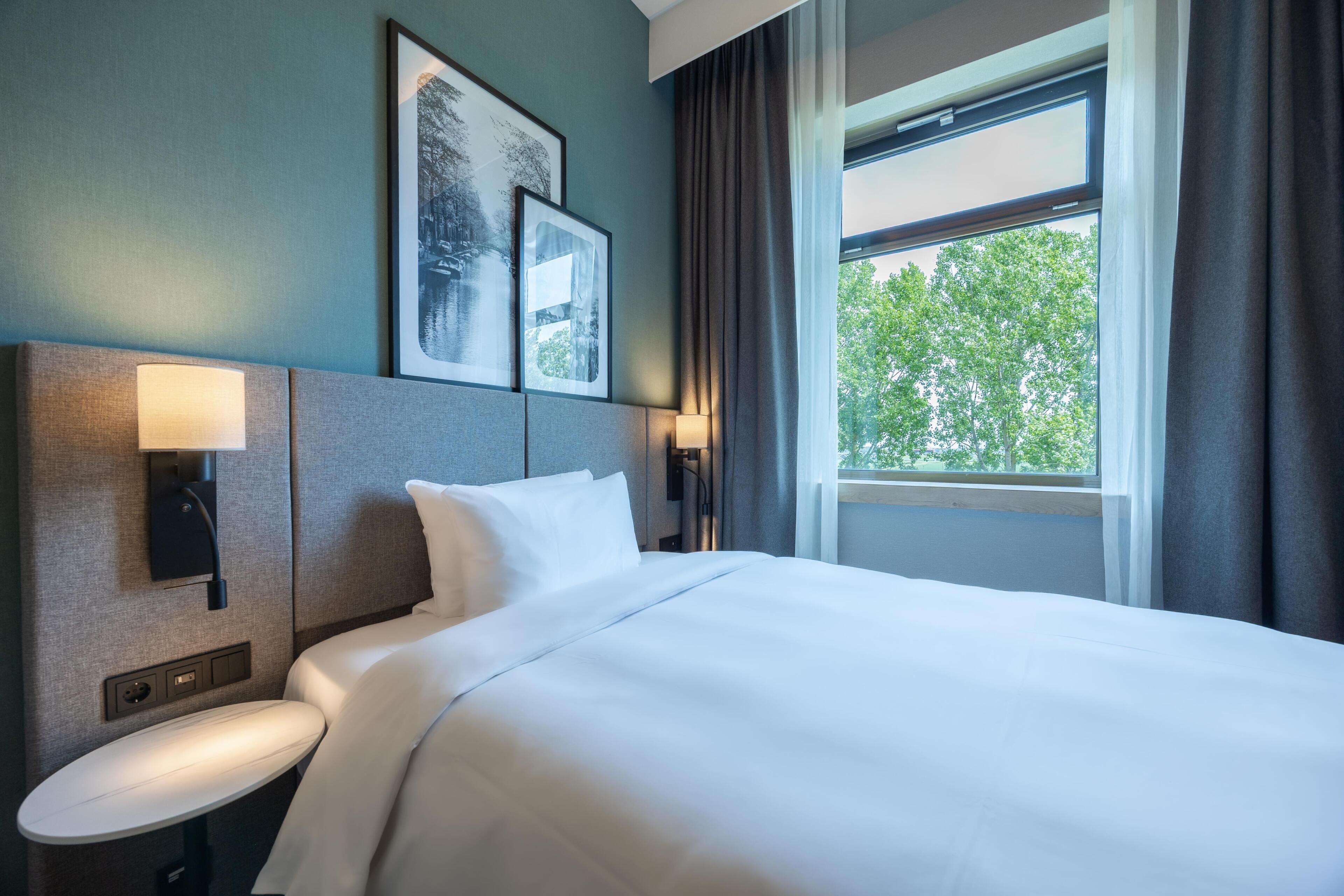 Habitación de hotel con una cama grande, dos lámparas de pared, una mesita y una ventana con cortinas.