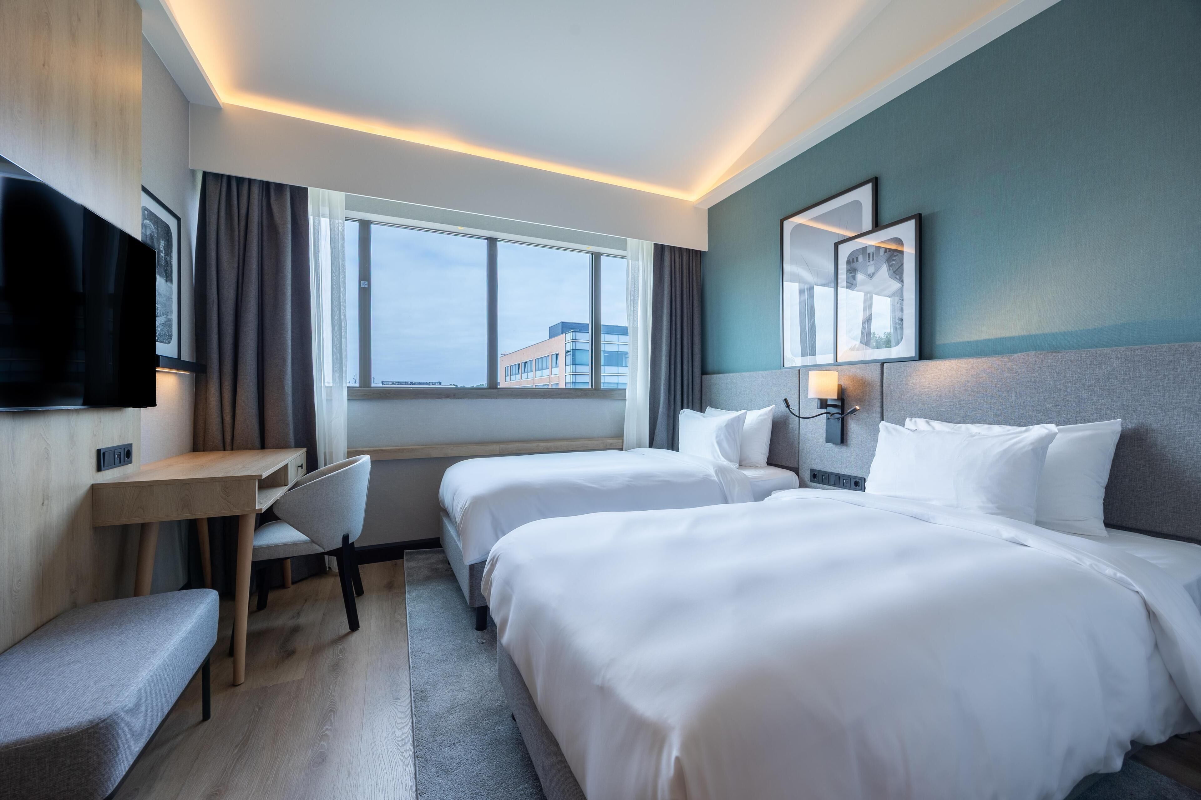 Ein Hotelzimmer mit zwei Betten, einem Schreibtisch, einem Stuhl, einem Fernseher und einem Fenster mit Blick auf Gebäude.