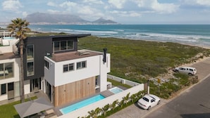 A modern two-story building with a rooftop pool, a view of the ocean, and a mountain in the distance.