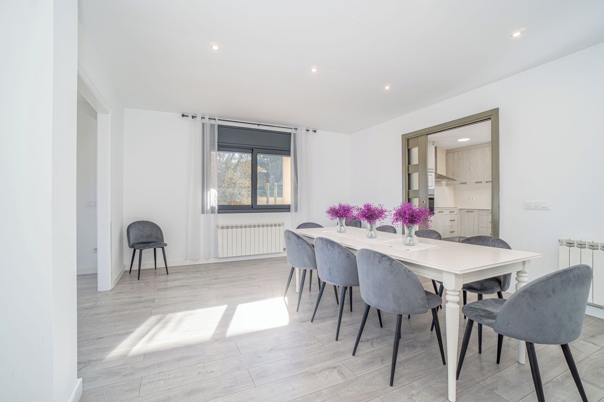 A modern dining area with a white table, grey chairs, and a window with natural light.