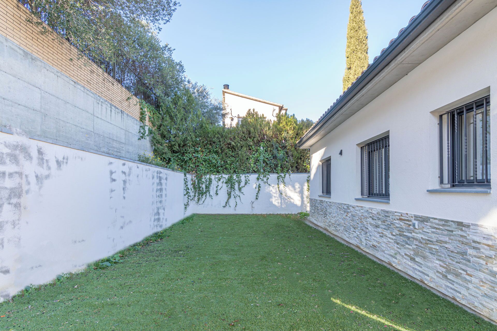 Un patio con césped, una pared blanca con detalles de piedra y un edificio con ventanas y una chimenea.